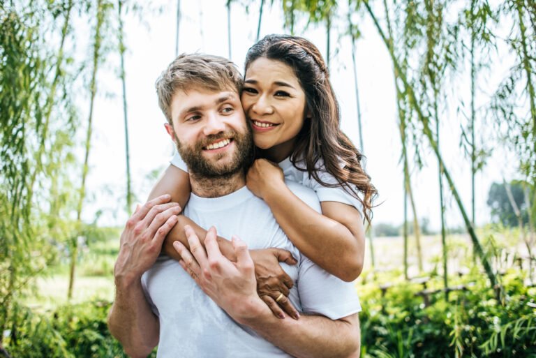 Smiling woman hugging a bearded man from behind as they stand outdoors surrounded by greenery.