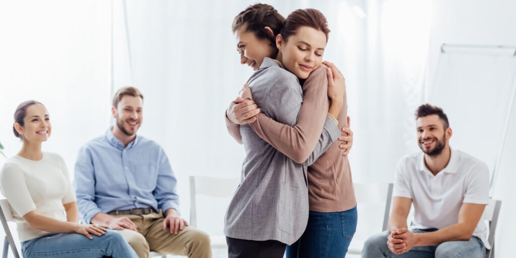 Two women hugging emotionally in a group therapy session while other support group members smile and watch with encouragement.