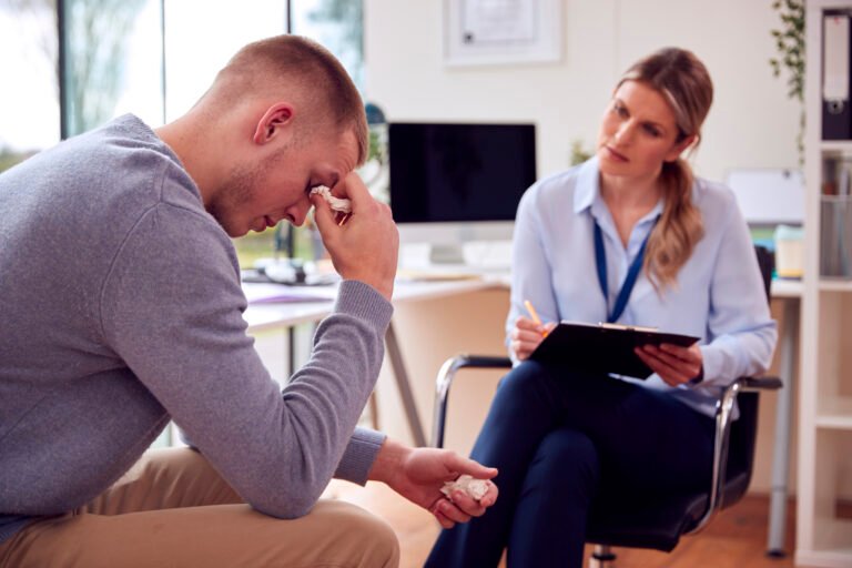 Distressed young man wiping tears during a therapy session with a female psychologist taking notes and listening attentively.