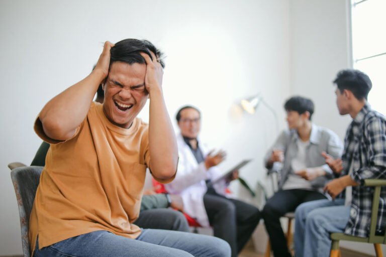 Young man screaming and holding his head in distress during a group therapy session with other participants and a counselor in the background.